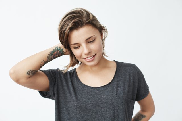 Shy young pretty tattooed girl smiling looking down over white background.
