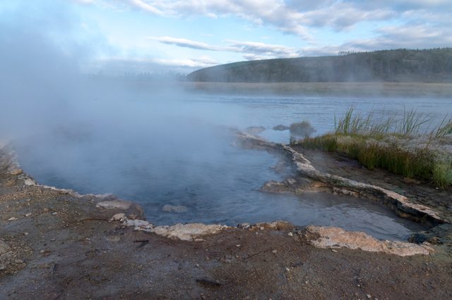 Steamy river water and muddy shoreline with green grass under blue sky with white fluffy clouds.
