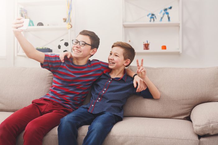 Technology, friendship and communication concept - two cute teenage boys taking selfie while posing with smile to phone and sitting on the couch in living room at home