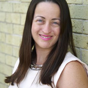 Woman smiling against a yellow brick wall, wearing a sleeveless white top and a beaded necklace.