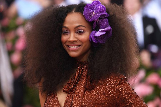 Person smiling, wearing sequined outfit with large purple flowers in hair, amidst a blurred background of people and floral decorations.