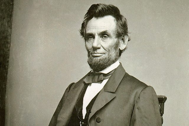 A seated man with a beard is wearing a suit with a bow tie, against a plain backdrop.