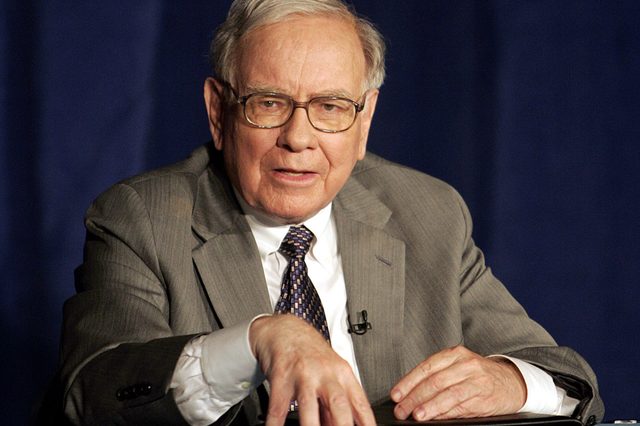An elderly man sits at a table, gesturing with his hand; he wears a suit and tie, with a dark blue background.