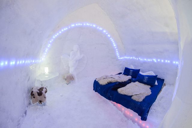 Ice bed covered in fur and pillows, set within an illuminated igloo room, featuring a snow sculpture and ambient blue lights.