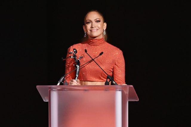 A person in a glittery red outfit holds a statuette, standing behind a translucent podium with microphones, against a dark background.