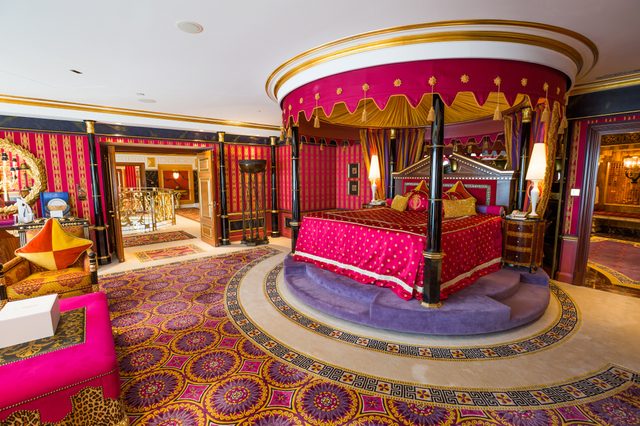 Luxurious bedroom with round bed, ornate patterns, vibrant red and gold colors, surrounded by rich decor and lavish furnishings in an opulent setting.