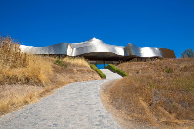 A metallic, wavy-roofed building sits on a hill, with a stone pathway leading through dry grass under a bright blue sky.