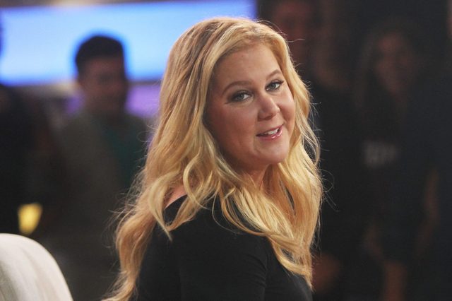 Person smiling, facing sideways in a studio with people blurred in the background, wearing a black top.