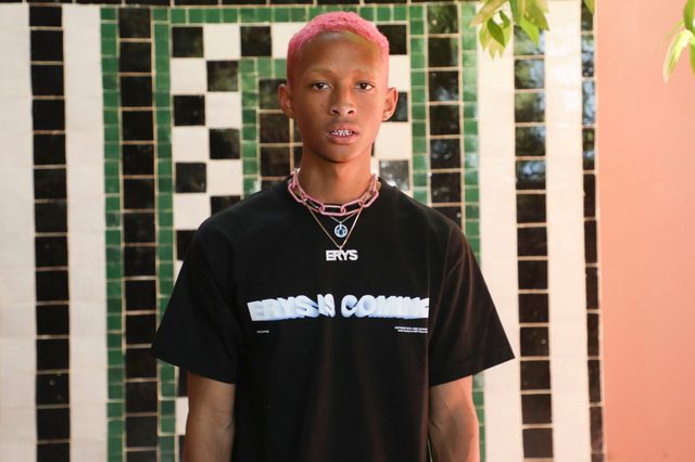Individual stands outside wearing a black shirt with text "ERYS IS COMING." Background features a geometric green and white tiled wall, with foliage above.