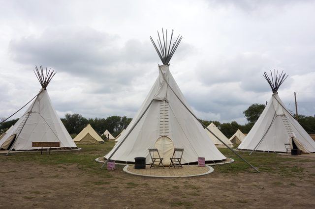 Tipis stand on grassy field, chairs positioned outside, surrounded by cloudy sky and additional tents in the background.