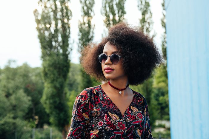 Thoughtful pretty afro-american woman in colorful blouse, wearing sunglasses, looking away. Outdoors.