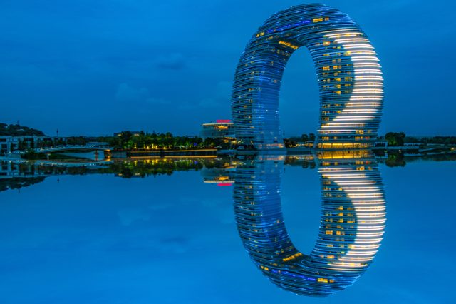 Circular building reflects in calm water, illuminated at night. Surrounded by trees and distant structures under a deep blue sky.