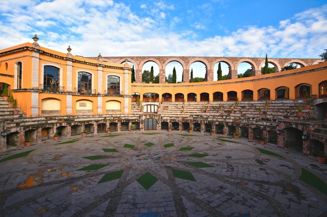 Stone amphitheater sits in front of a peach building with arched windows, surrounded by trees and blue sky.