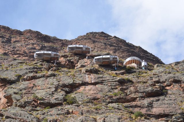 Pods hang on a rocky cliffside, overlooking a mountainous landscape with clear skies above.