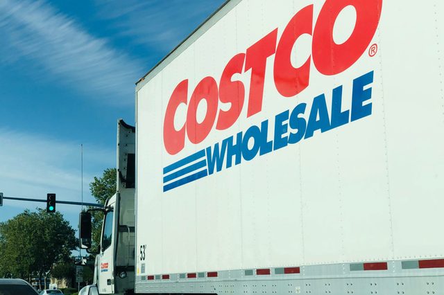 White delivery truck displaying "COSTCO WHOLESALE" logo, driving on a tree-lined road beneath a clear blue sky.