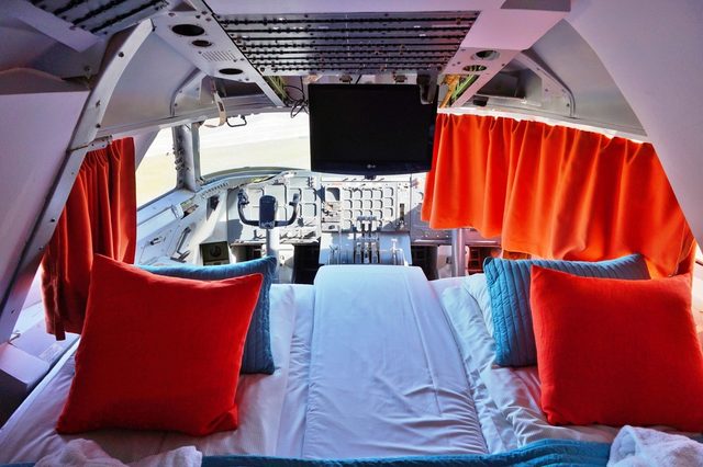 Bed sits in airplane cockpit with orange curtains and blue cushions, blending aviation elements into a cozy sleeping area.