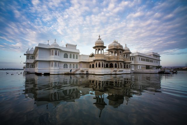 White palace reflecting on lake, surrounded by calm water and a partly cloudy sky, creating a serene and majestic atmosphere.