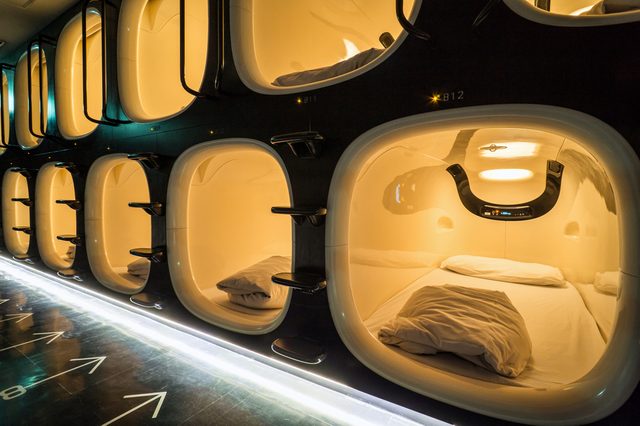 Pods arranged vertically are lit warmly, showing compact beds inside. They are in a futuristic sleeping area with glowing floor arrows.