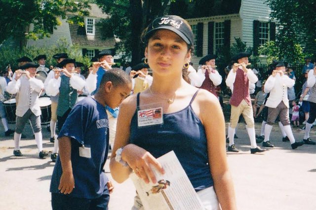 Girl holding a paper, standing in front of a marching band in colonial attire, outdoors with trees and historic buildings in the background.