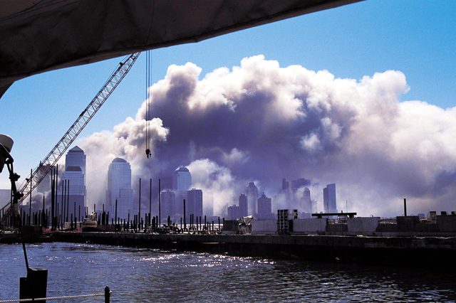Smoke billows above skyscrapers, framed by a construction crane, with water and docks in the foreground under a clear blue sky.