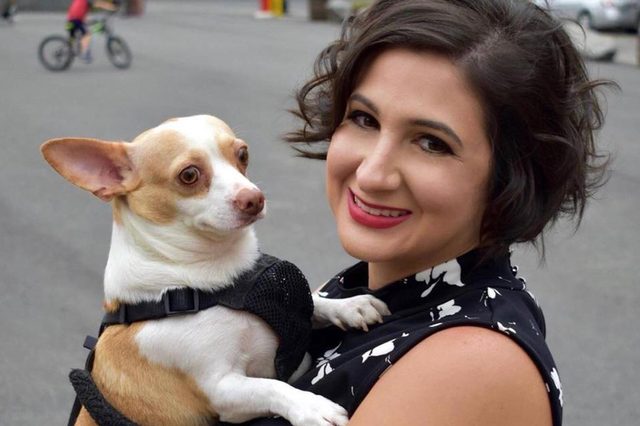 A woman holds a small dog, both looking at the camera, on an outdoor street with blurred cyclist in the background.
