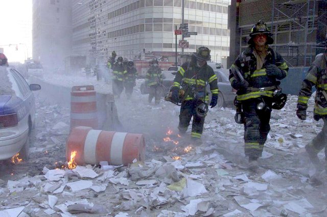Firefighters walk through debris and smoke, amidst scattered papers and small fires, on a city street flanked by tall buildings.