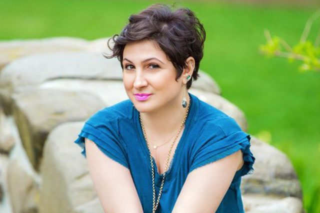 A person with short hair smiles, sitting against a stone wall, wearing a blue shirt; greenery fills the background.