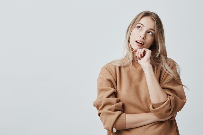Portrait of pensive fair-haired dreamy hipster girl imagines something in her mind, looks upwards, being deep in thoughts, touching chin. Attractive young blonde female dreams about weekends