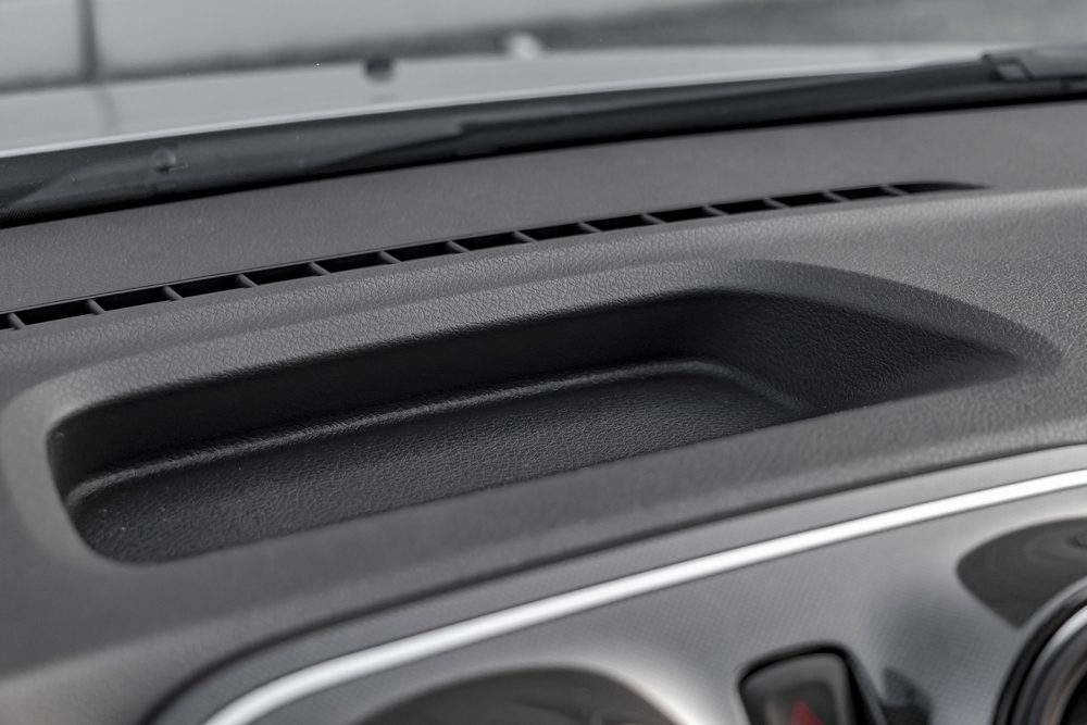 Dashboard storage compartment sits unused, surrounded by textured material, near a windshield and car controls.