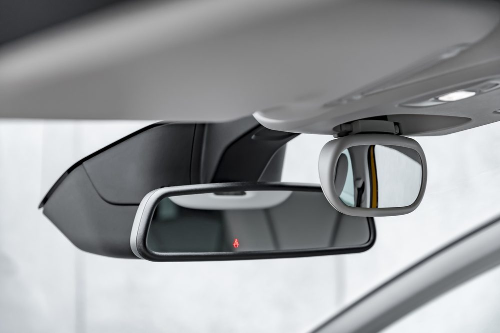 Rearview mirror displays red warning light inside a car, with a dual sun visor visible above and white car interior surrounding.