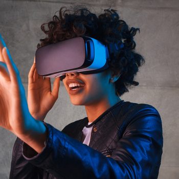 Person wearing virtual reality headset, reaching out with hand in a concrete-walled room.