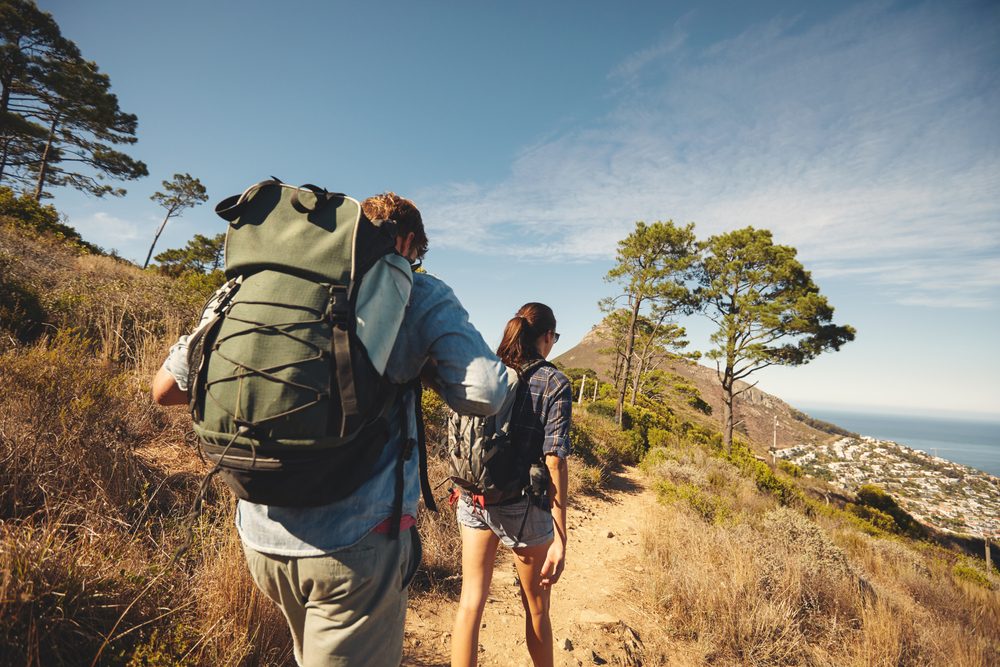 Two hikers walk on a dirt trail surrounded by trees and coastal scenery under a clear blue sky.