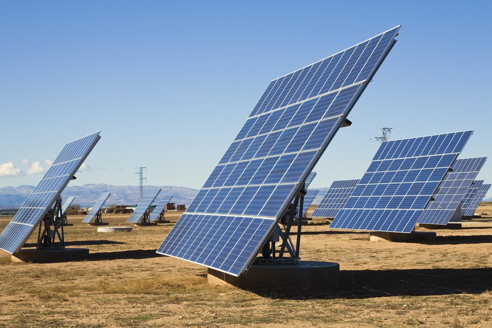 Solar panels stand capturing sunlight, arranged in rows on a dry, grassy landscape with distant mountains under a clear blue sky.
