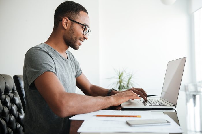 Happy african young man dressed in gey t-shirt and wearing eyeglasses using laptop and sitting at the table.