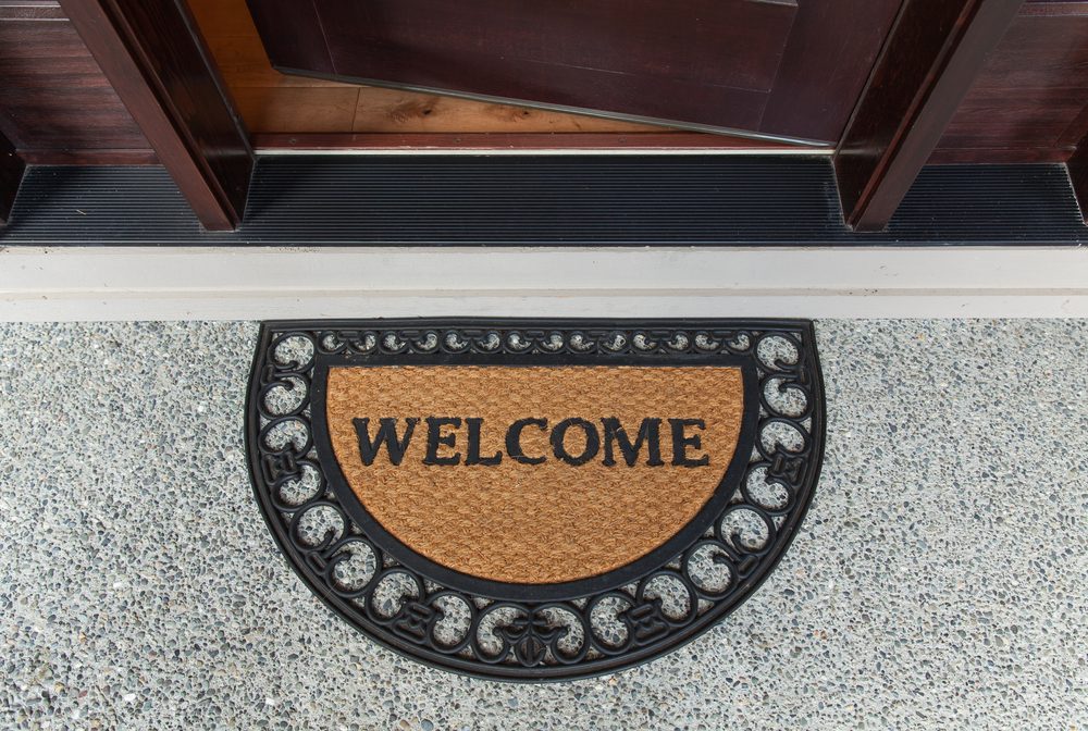 Doormat displays "WELCOME" at a doorstep, resting on speckled concrete, surrounded by a decorative black border.