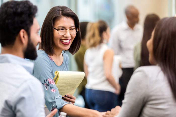 A cheerful young businesswoman stands with an unrecognizable coworker in a crowded office building and smiles as she shakes hands with her new unrecognizable client.