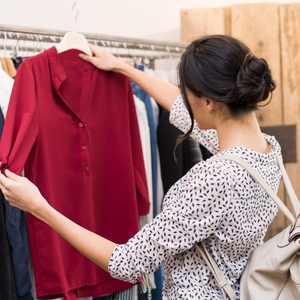 Woman examines a red blouse in a clothing store, surrounded by various clothes on hangers.