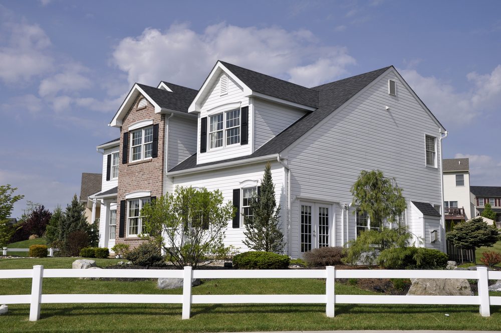A two-story house stands surrounded by a white fence, with manicured shrubs and a blue sky overhead.