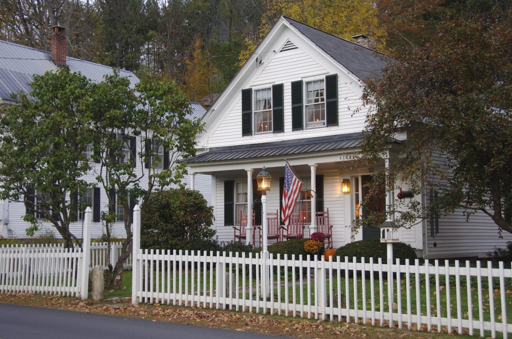 White house displays American flag, surrounded by a white picket fence and trees, with autumnal foliage in the background. Porch has red chairs.