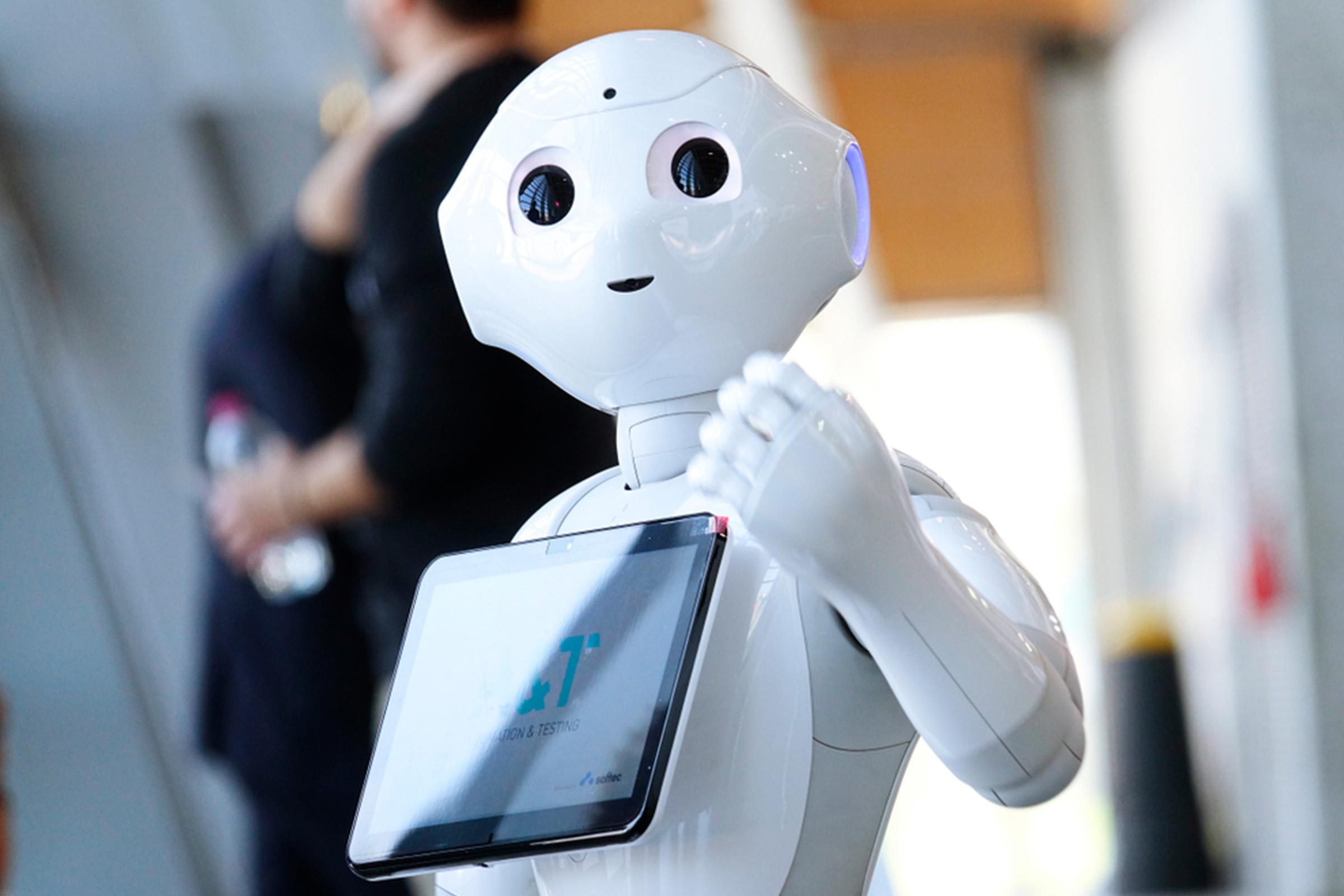 A humanoid robot gestures with a screen on its chest, standing indoors among people, displaying "TESTING" text.