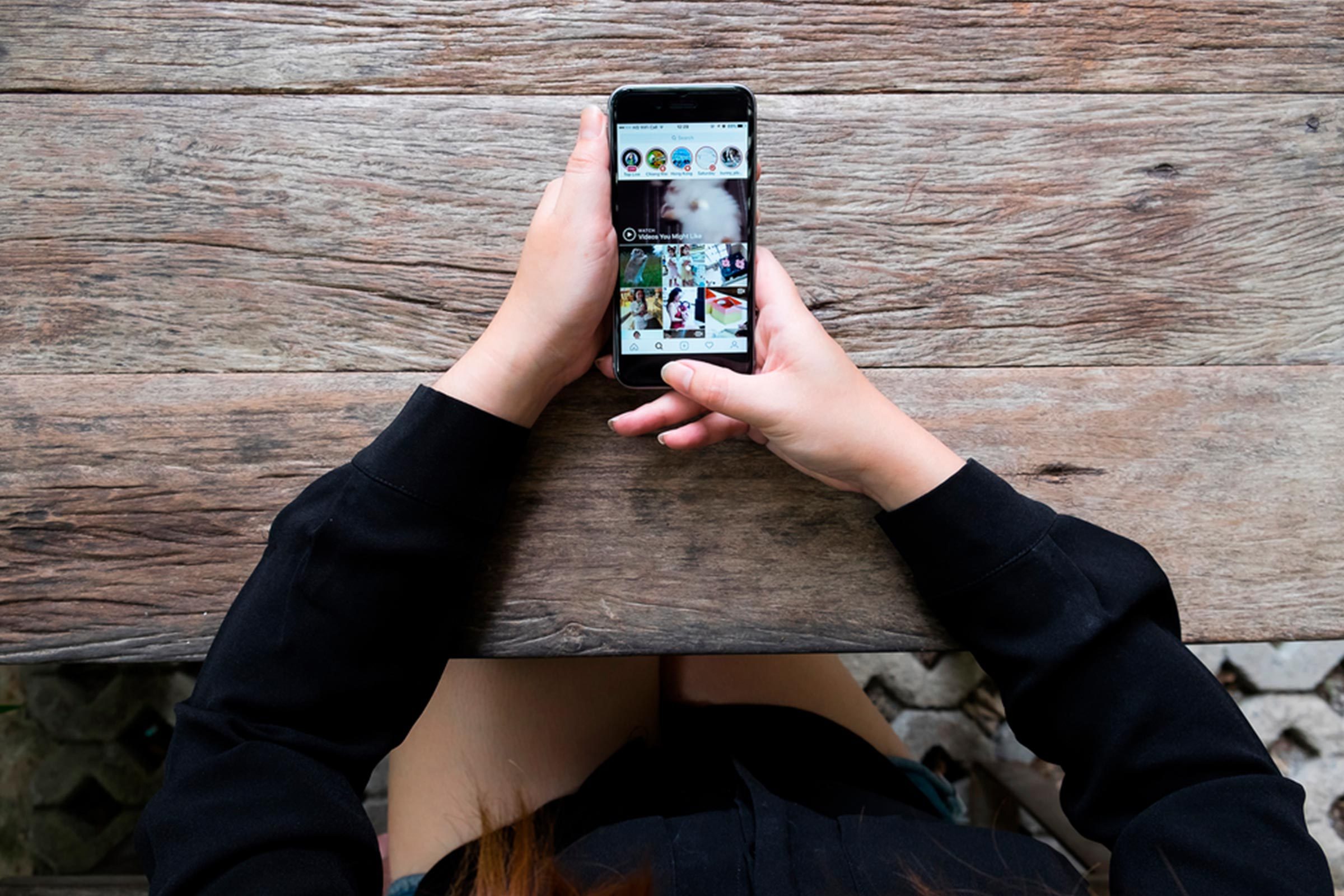 Hands holding a smartphone, browsing social media on a wooden table with a blurred background.