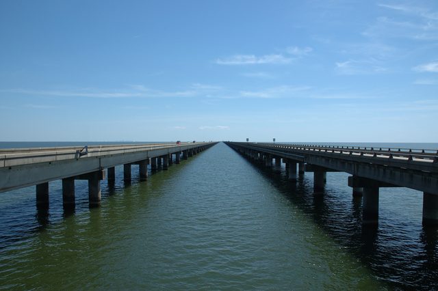 Two long bridges stretch across a body of water under a clear blue sky, converging towards the horizon.