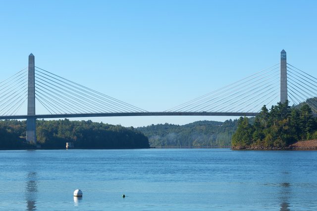 A suspension bridge stretches over a calm river, surrounded by forested hills under a clear blue sky.