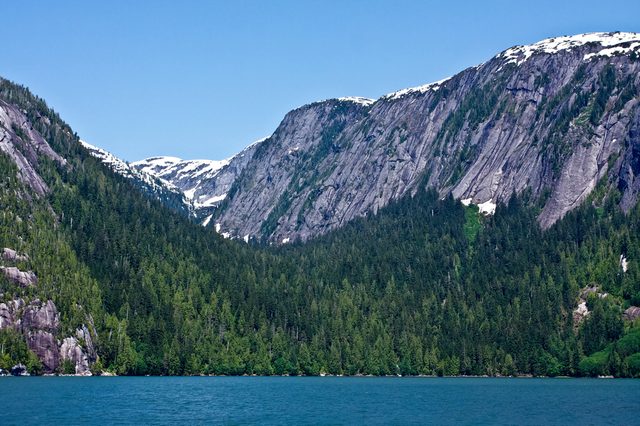 Water reflects mountains covered in dense green forest with patches of snow under a clear blue sky.