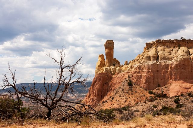 Rock formation towers amidst arid terrain, surrounded by dry brush and partially cloudy sky.