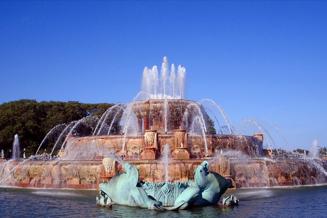 A large, ornate fountain sprays water in tiered arcs, surrounded by sculptures, with a clear blue sky and trees in the background.