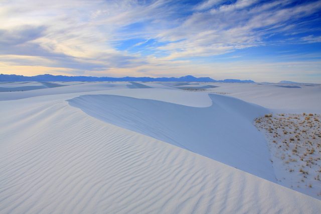 White sand dunes stretch across the landscape under a vast blue sky with clouds, surrounded by distant mountains and patches of sparse vegetation.