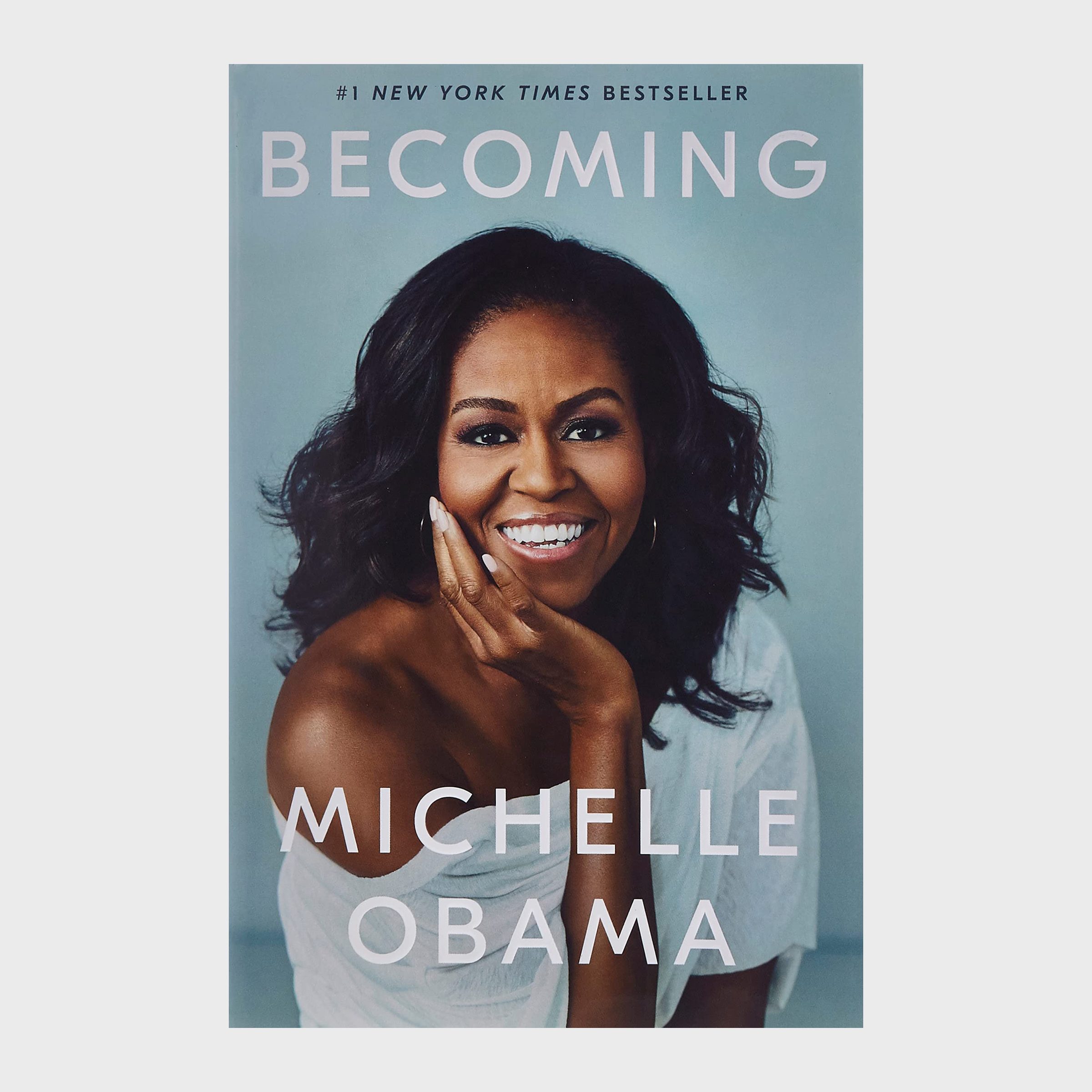 A woman smiles while resting her chin on her hand; text reads, â€œ#1 New York Times Bestseller, Becoming, Michelle Obama.â€ Background is light blue.