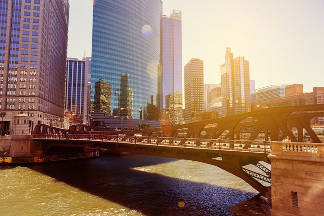 Bridge spans a river under sunlight, surrounded by tall skyscrapers in an urban skyline.