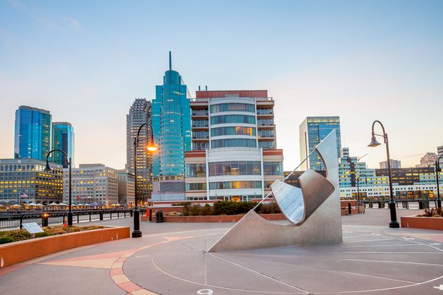 Metal sculpture stands on a city plaza, surrounded by modern high-rise buildings and lampposts, under a clear sky at dusk.